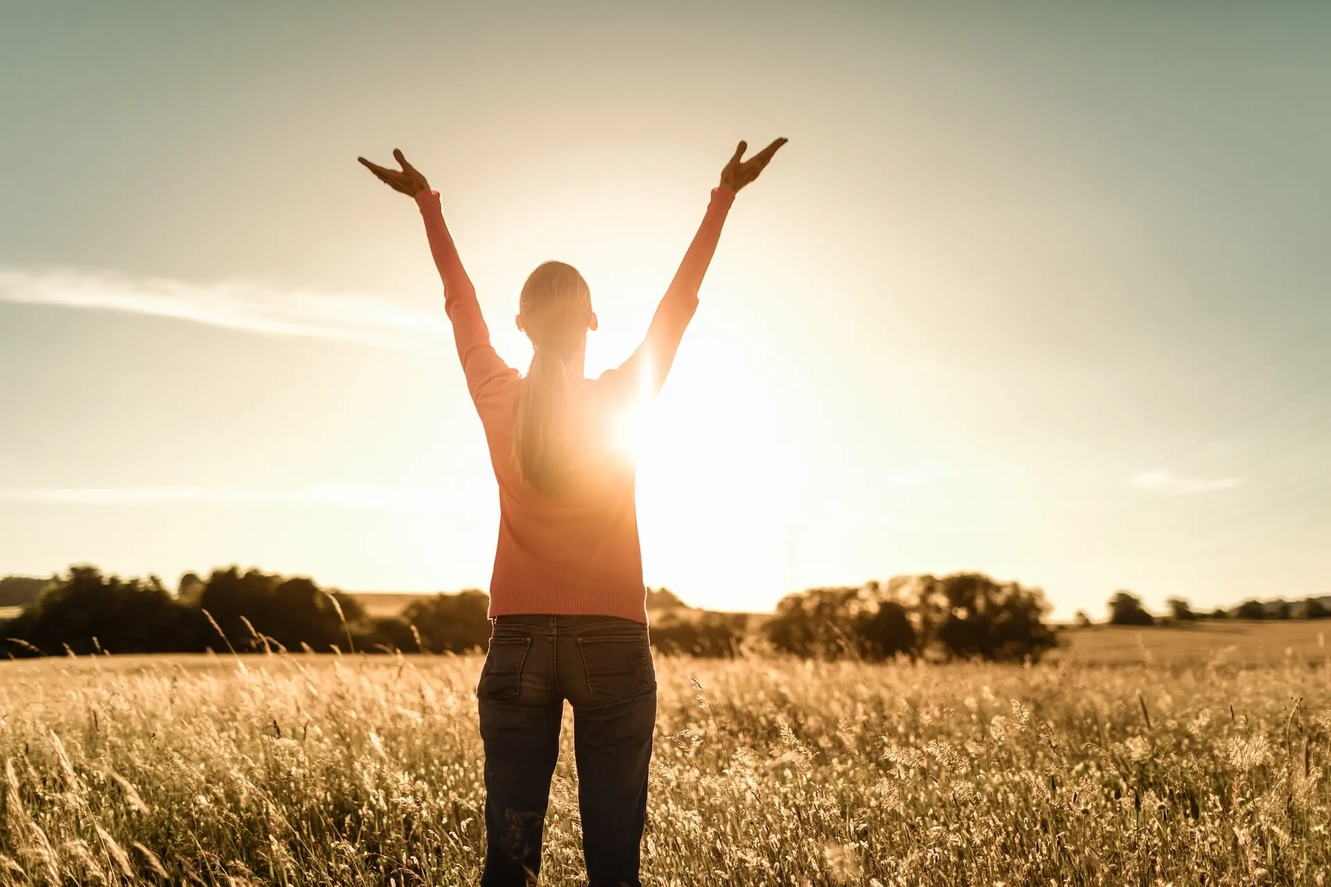 Person standing in tall grass with arms raised during golden sunrise